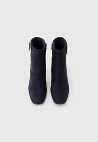 Une paire de bottines en daim noir avec des bouts carrés et des détails en boucle dorée sur un fond gris clair.