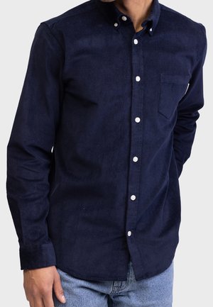 Homme portant une chemise en velours côtelé bleu marine avec des boutons blancs et une poche poitrine, associée à un jean bleu clair.