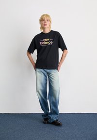 Asmuo su trumpais šviesiais plaukais, vilkintis juodą New Balance marškinėlį, šviesiai mėlynas išblukusias džinsus ir juodus batus, stovi ant mėlyno kilimo.
