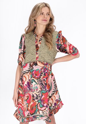 Blondynka w kolorowej sukience w kwiaty z oliwkowozieloną koronką na szydełku i kolczykami z koralików, pozująca z ręką na biodrze.