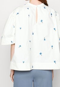 White cotton blouse with short, puffed sleeves. Features blue floral embroidery and a back keyhole opening with a button closure.
