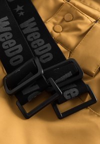Svart nylonbälte med dubbla fyrkantiga spännen, med ordet "WeeDo" tryckt i grått. Mot en texturerad gul tygbakgrund.