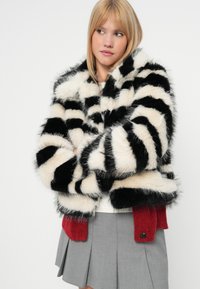 Suknja iz umetnega krzna s črnimi in belimi črtami, ki se ponaša s prijetno teksturo, širokimi rokavi ter rdečim pletenim detajlom na robu.