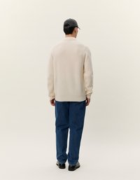 Uomo di spalle che indossa un maglione color crema, jeans blu, scarpe nere e un cappellino da baseball nero su uno sfondo chiaro e semplice.