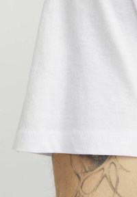 Witte katoenen t-shirt met een rechte zoom, korte mouwen en een gladde textuur, die gedeeltelijk een arm bedekt met een zichtbare tatoeage.