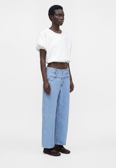 Personne debout portant un haut blanc à manches courtes, un jean bleu taille haute à jambes larges et des chaussures plates marron foncé sur fond blanc.