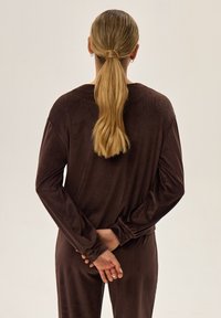 Brązowa, teksturowana bluzka z długim rękawem o luźnym kroju i bufkowatych rękawach, widok z tyłu. Włosy ułożone w niskiego kucyka.