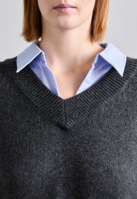 Maglione grigio in lana con scollo a V profondo su una camicia colletto azzurro. La texture è lavorata a maglia e morbida, con polsini e orlo ribattuti.
