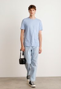Camiseta de manga curta azul clara, jeans de denim claro folgados, mochila a tiracolo preta e sapatilhas preto e branco. Design simples e ajuste casual.