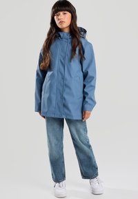 Giacca impermeabile azzurro chiaro con cappuccio, bottoni a pressione e tasche laterali, abbinata a jeans blu a vestibilità rilassata e sneaker bianche.