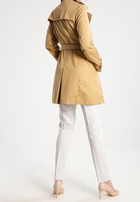 Cappotto trench khaki con cintura in vita, spacco posteriore e ampio colletto, abbinato a pantaloni chiari in tessuto texturizzato e tacchi nudi. Finitura liscia.