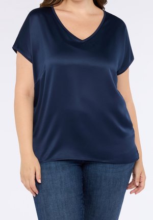 Donna che indossa una camicetta di raso blu navy a maniche corte con scollo a V e jeans blu, mostrata dal collo ai fianchi su uno sfondo neutro.