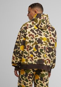 Felpa camouflage in beige, marrone e verde con motivi di faccine sorridenti. Presenta un grande cappuccio e una vestibilità ampia con polsini ed orlo elasticizzati.