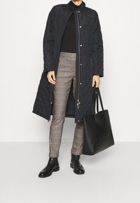 Abrigo largo de tejido acolchado en color azul marino, suéter negro de cuello alto, pantalones de cuadros, botas de tobillo de cuero negro y un gran bolso de mano negro. Cuenta con cierres de cremallera y a presión.