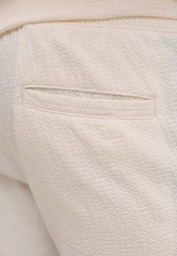 Gros plan d’un pantalon en tissu beige texturé montrant une poche arrière horizontale avec un motif discret et des détails de couture.
