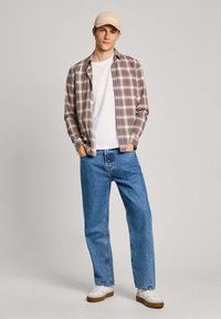 Camisa de cuadros en marrón, azul marino y blanco; camiseta blanca; jeans azules; gorra beige con logo; zapatillas blancas con suela marrón. Atuendo casual.