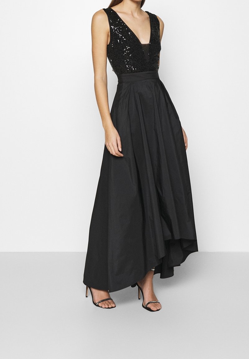 Femme portant une robe noire sans manches avec un corsage en V profond orné de sequins et une jupe longue asymétrique, associée à des sandales noires à talons hauts.