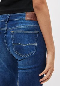 Niebieskie jeansy z denimu o średnim stanie, z brązową skórzaną naszywką na pasku i kontrastowymi przeszyciami na tylnych kieszeniach.