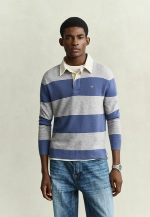 Junger Mann trägt ein blau-grau gestreiftes Langarm-Poloshirt, hellblaue Jeans und eine silberne Armbanduhr und steht vor einem schlichten Hintergrund.