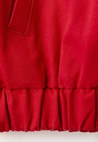 Prim-plan cu material roșu cu tiv elastic adunat și detaliu de cusătură vizibil pe o textură netedă, țesută.