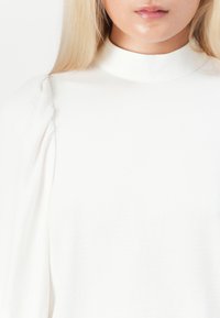 Woman wearing a white high-neck blouse with puffed sleeves, showing only from mid-chest to lips on a white background.