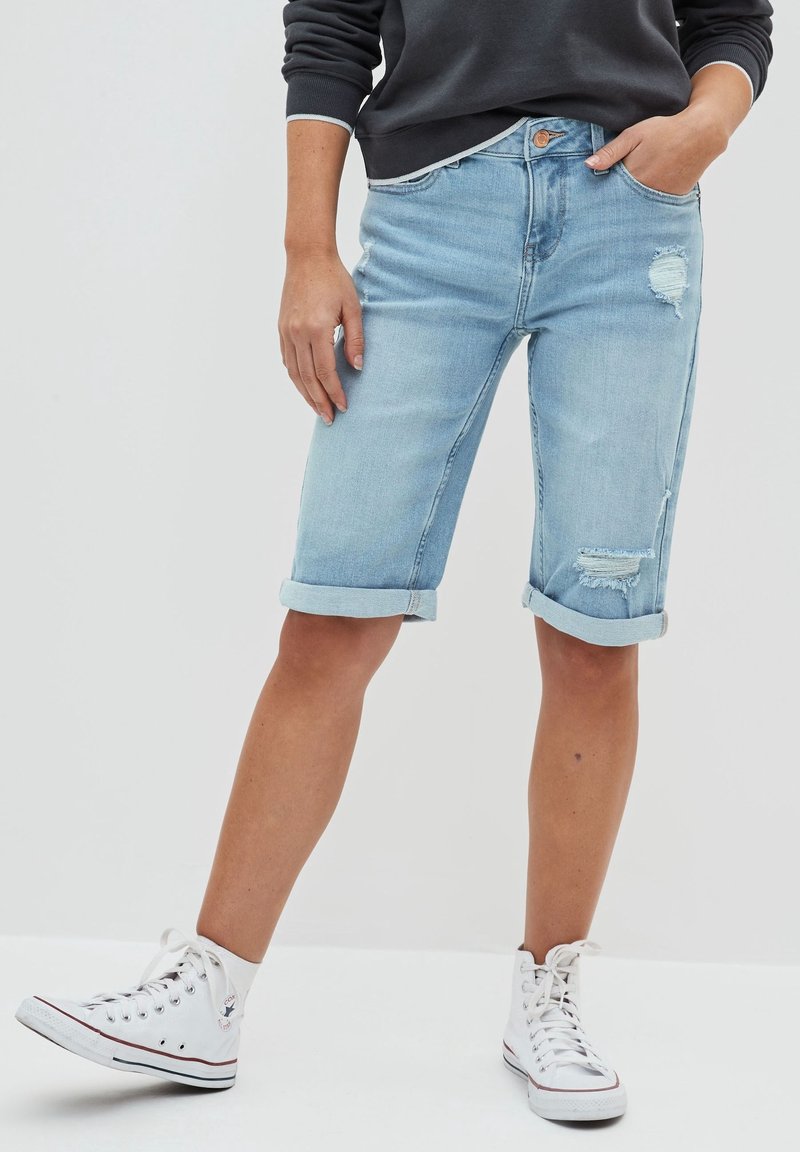 Personne portant un short en denim bleu clair retroussé avec des détails usés, un sweat-shirt noir et des baskets montantes blanches, debout devant un fond uni.