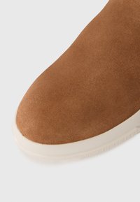 Nærbilde av en brun semsket skobot med en hvit gummisåle på en hvit bakgrunn.