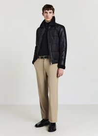Veste noire en cuir matelassé avec un col texturé, associée à un pantalon beige et des chaussures noires, présentant un design épuré.