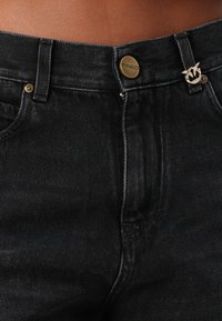 Crne traperice od denima s visokim strukom, prednjim džepovima, zlatnim detaljima i oznakom brenda na dugmetu sa logotipom na pojasu.