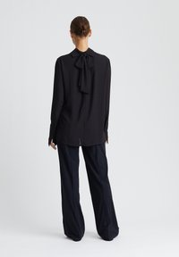 Chemise noire à manches longues avec un détail nœud papillon au col, en tissu lisse, avec des poignets boutonnés, associée à un pantalon noir à jambes larges.