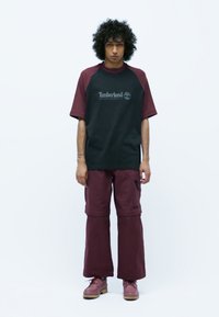 Junger Mann mit lockigem Haar, der ein schwarzes und burgunderfarbenes Timberland-T-Shirt, burgunderfarbene Lagenhosen und rosafarbene Stiefel trägt, steht vor einem weißen Hintergrund.