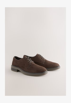 Brune ruskindssko med glat tekstur, rund snude, fem snørehuller og en kontrasterende syningsdetalje på sålen.