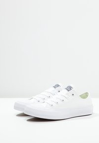 Las zapatillas blancas de lona presentan un diseño de caña baja, punta redonda y un cierre clásico con cordones. Los acentos incluyen un parche de logo azul marino.