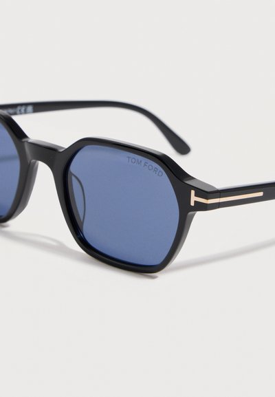 Čierne osemuholníkové slnečné okuliare s tmavomodrými šošovkami a zlatými "T" ozdobami na straniciach, s logom "TOM FORD" na šošovke.