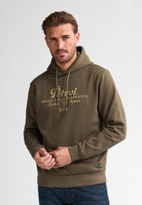 Olivově zelená mikina s kapucí a stahovací šňůrkou, s texturovaným logem a grafikou ve světlé béžové barvě. Hribkované manžety a lem, s nášivkou na spodní části.