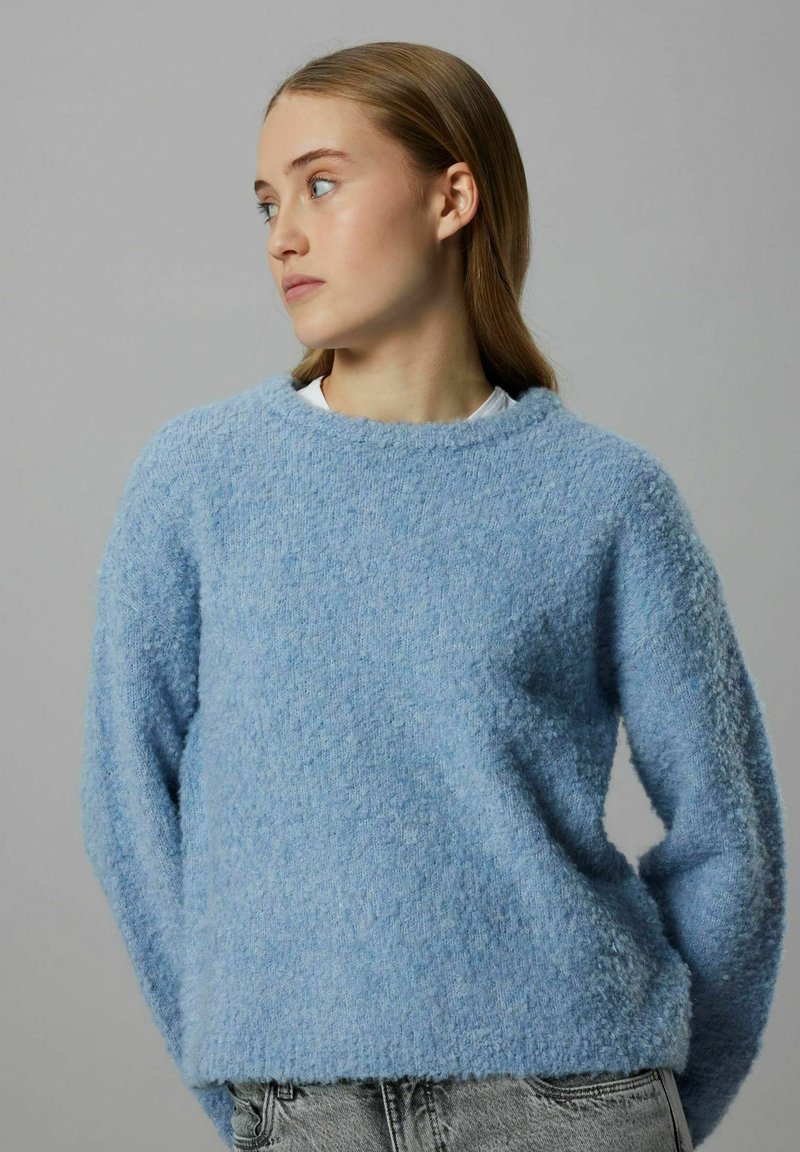 Gaiši zils pūkains džemperis ar apaļu apkakli, garām piedurknēm un teksturētu audumu. Valkāts pār baltu topu, kombinēts ar pelēkām džinsu biksēm.