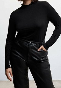 Femme portant un haut noir moulant à manches longues rentré dans un pantalon en cuir noir taille haute, une main dans la poche, sur fond uni.