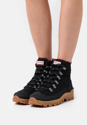 Bottes de randonnée noires avec une semelle en caoutchouc, tige en tissu, œillets en métal et accents rouges contrastants sur le talon et l'étiquette. Design texturé et robuste.