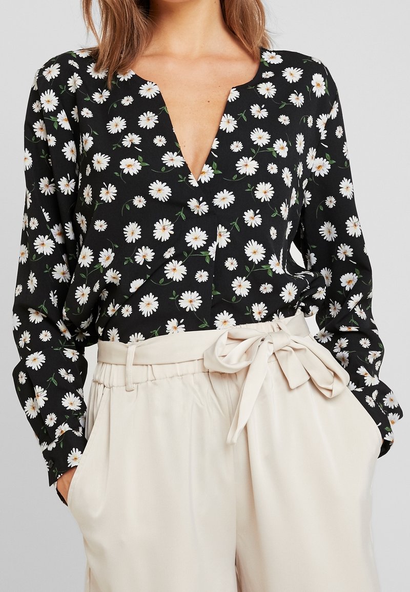 Blouse noire à motif floral avec marguerites blanches, col en V plongeant, manches longues, associée à un pantalon en lin beige avec taille nouée et poches avant.