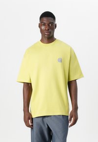 T-shirt en coton vert lime surdimensionné avec un col rond côtelé et un petit logo violet sur la poitrine à gauche. Manches courtes, coupe décontractée.