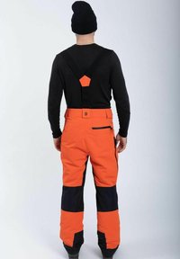 Oranssit vedenpitävät haalareita, joissa on mustia yksityiskohtia, säädettävät hihnat ja vetoketjulliset taskut. Sileä rakenne, kestävä materiaali ja tiukka istuvuus.