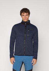 Jachetă din fleece de marină cu tricot texturat, guler înalt și accente negre. Are un fermoar frontal și un buzunar pe piept cu fermoar.