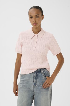 Frau trägt ein hellrosa kurzärmeliges, kabelgestricktes Poloshirt und ausgebleichte blaue Jeans, steht mit einer Hand in der Tasche.
