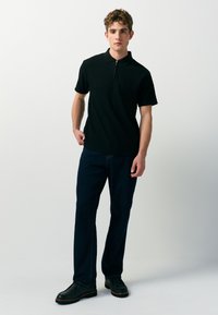 Schwarzes, geripptes Poloshirt mit einem Reißverschluss am Kragen, kombiniert mit dunkelblauen Jeans mit geradem Bein und schwarzen Schuhen. Das Modell steht vor einem neutralen Hintergrund.