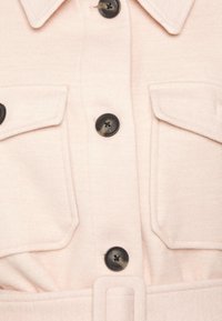Camisa rosa clara con tejido texturizado, dos bolsillos en el pecho y botones negros. Incluye un cinturón con hebilla rectangular.