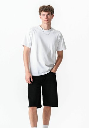 Jeune homme aux cheveux bouclés portant un t-shirt blanc, un short noir arrivant aux genoux, des chaussettes blanches et un collier fin en chaîne, debout avec une main dans la poche.