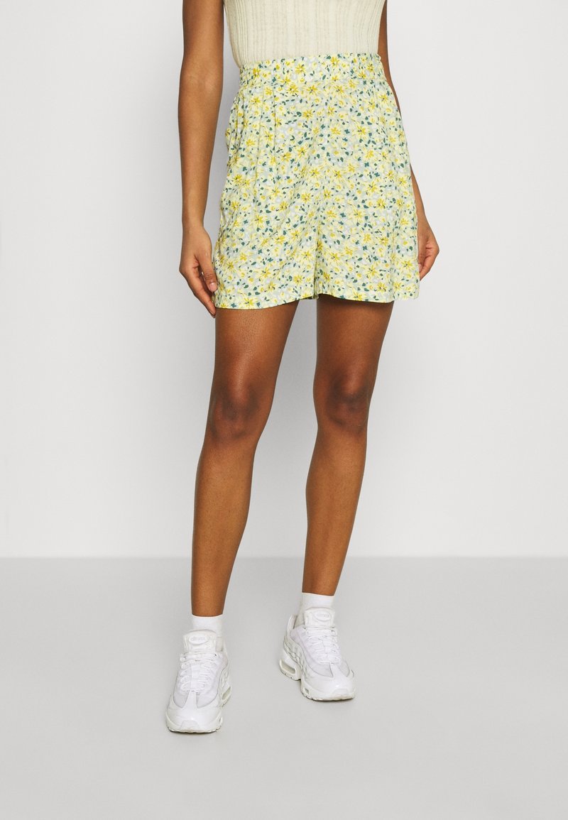 Personne portant un short taille haute fleuri jaune et blanc, des chaussettes blanches et des baskets blanches, debout devant un fond clair uni.