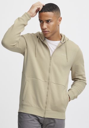 Beige zip-up hoodie with a hood, kangaroo pockets, and ribbed cuff detailing. Made of soft material with a smooth texture.