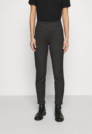 Personne portant un pantalon gris foncé ajusté avec des poches avant et une fermeture à bouton, associée à des bottines noires et un haut noir à manches courtes.