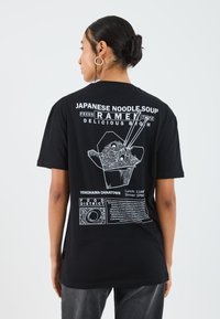 Svart t-skjorte med hvit grafisk trykk av ramen i en takeout-eske med tekst om mattilbudene i Yokohama Chinatown. Korte ermer.